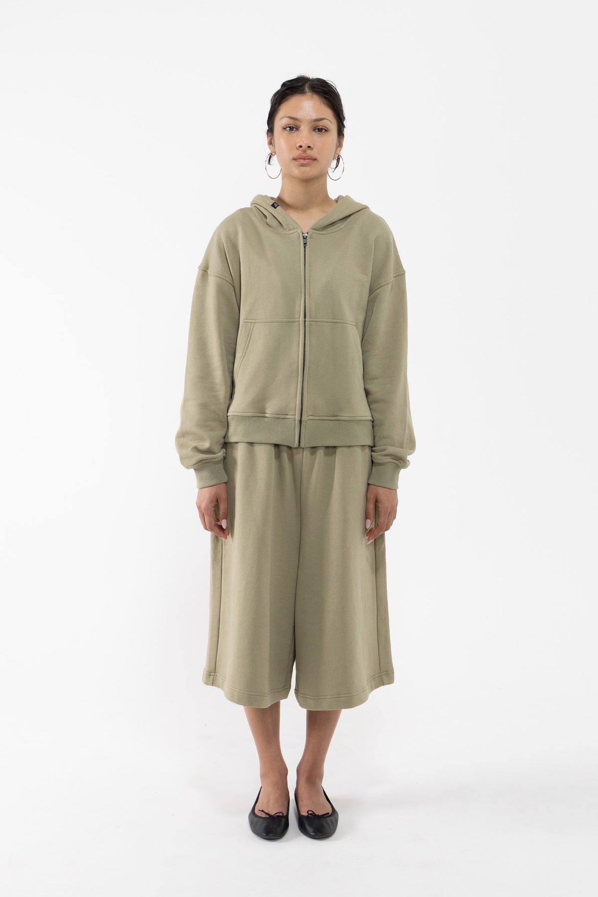 A dancer stands against a plain white studio background wearing a matching sage green two-piece lounge set consisting of a zip-up hooded sweatshirt and wide-leg cropped pants.