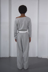 Person wearing a gray off-shoulder top and gray wide-leg pants against a white wall.