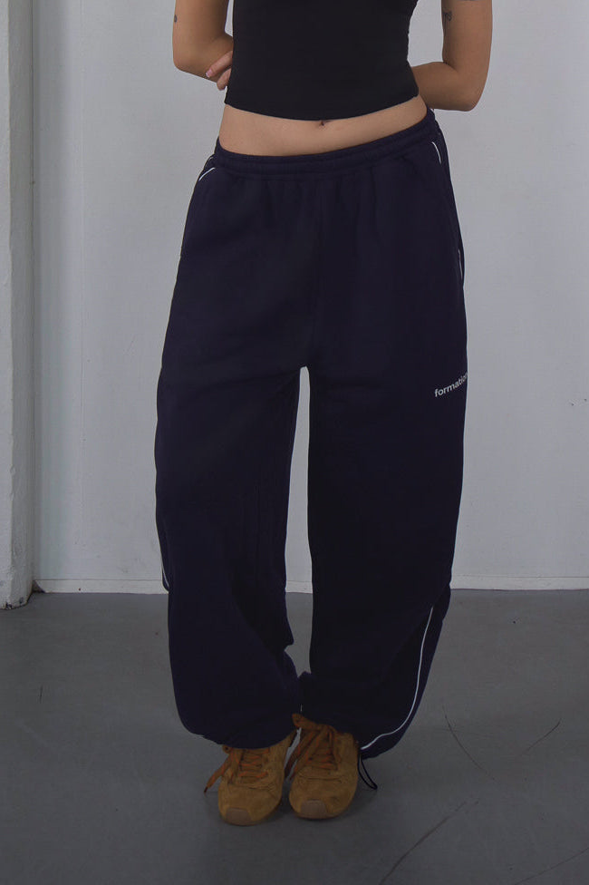 Woman wearing a black crop top and navy pants against a white wall.