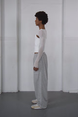 Person wearing a white long-sleeve top and gray pants standing against a white wall.