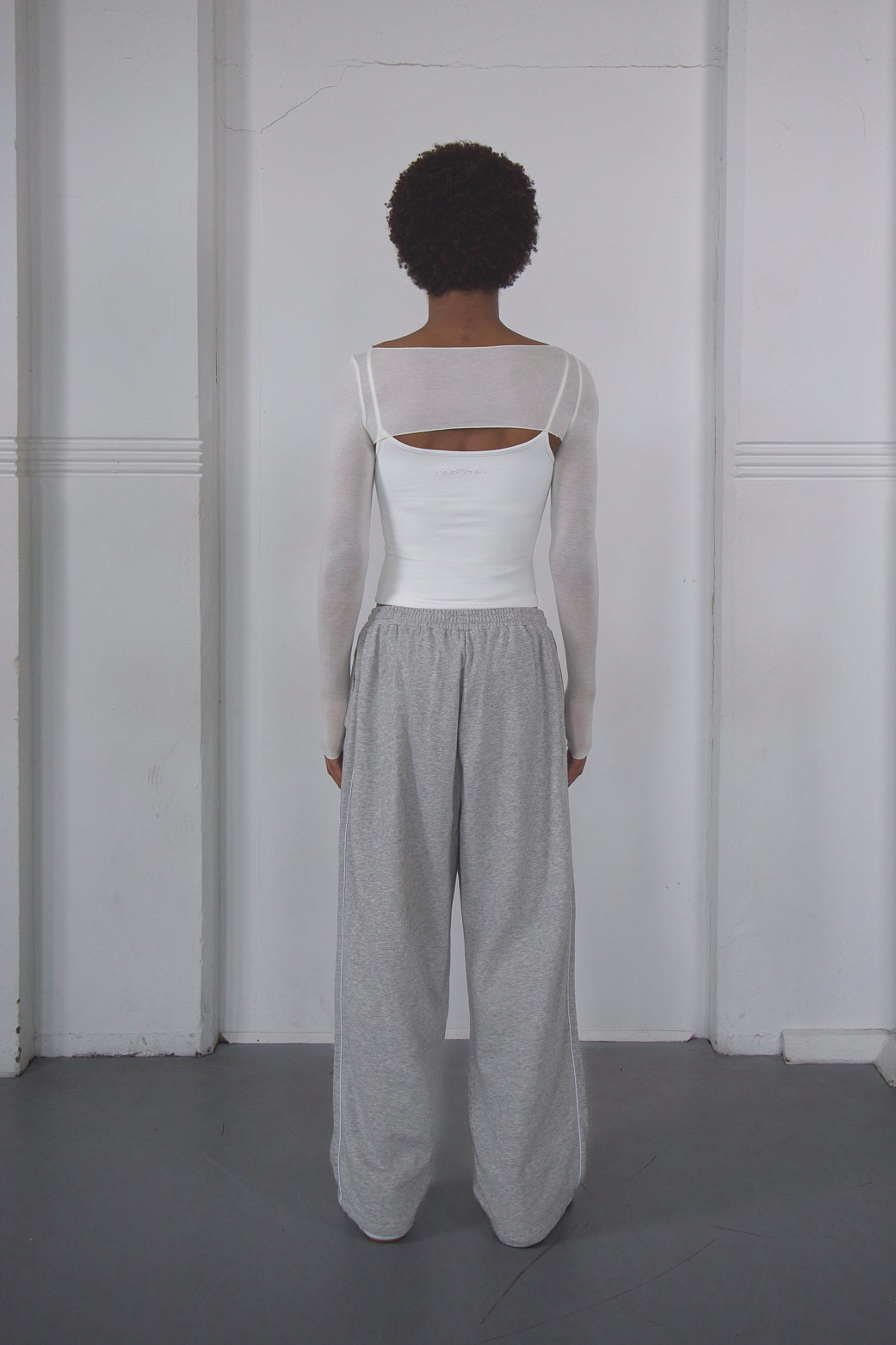 Person wearing a white top and grey pants against a white wall.