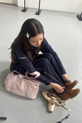 Woman sitting on the floor tying ballet shoes with a pink bag beside her. Wearing navy sweat set.