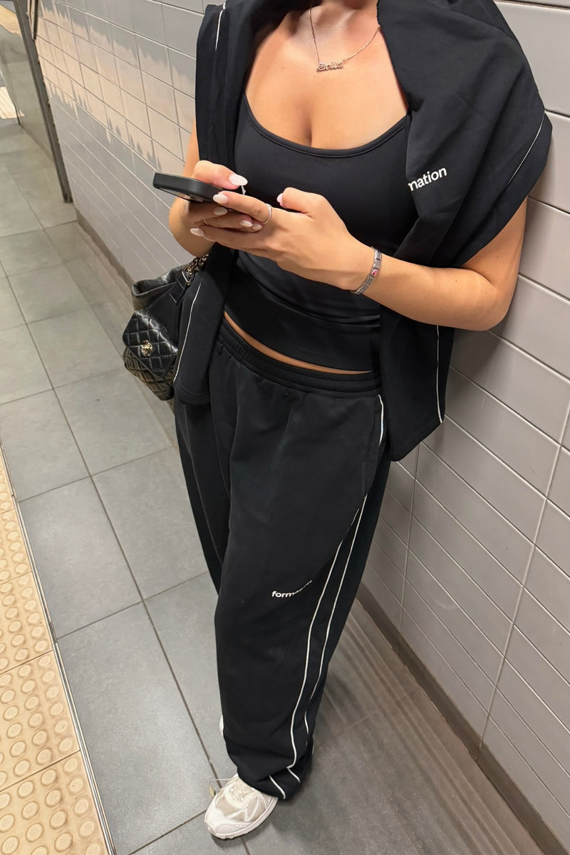 Person wearing a black tank top, sweatpants and coverup, standing against a tiled wall.