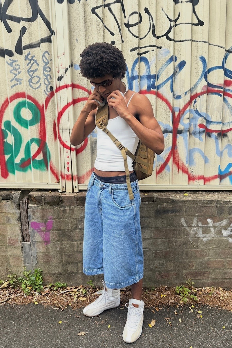 Person wearing a white tank top, blue jorts, and white sneakers in front of a graffiti-covered wall.
