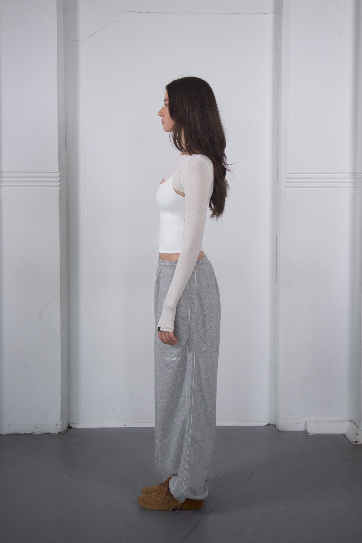 Woman wearing a white long-sleeve top and gray patterned pants standing against a white wall.