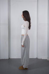 Woman wearing a white long-sleeve top and gray patterned pants standing against a white wall.